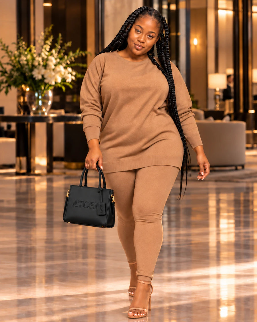 Model wearing a camel two-piece sweater top and leggings set with nude strappy heels, carrying a black ATORI tote through a hotel lobby.